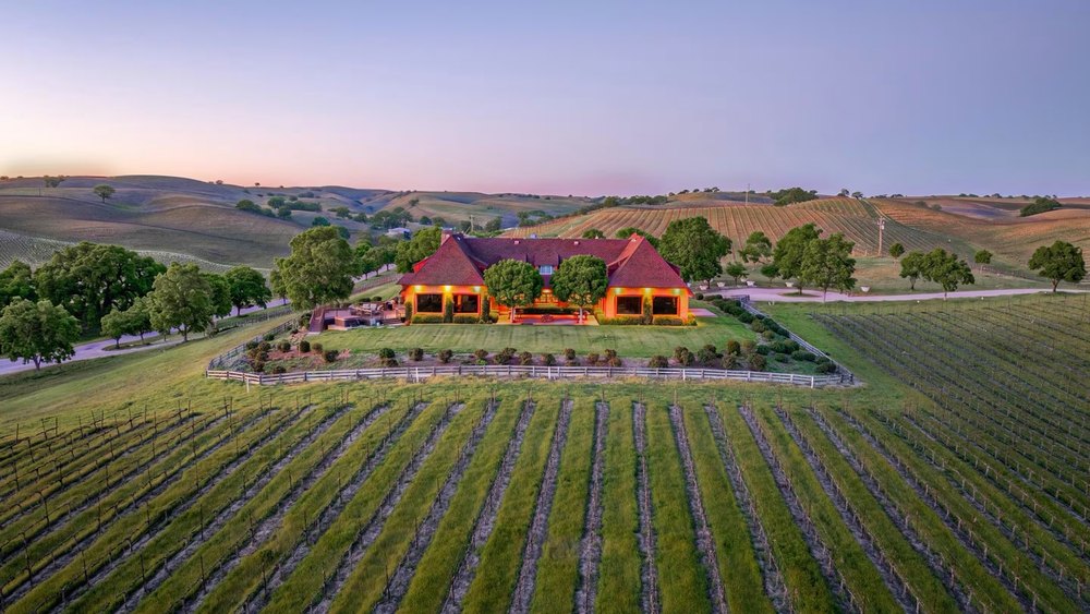 Aerial view of a sprawling vineyard estate at sunset, featuring a glowing ranch-style main house nestled among rolling green hills and neat rows of grapevines.