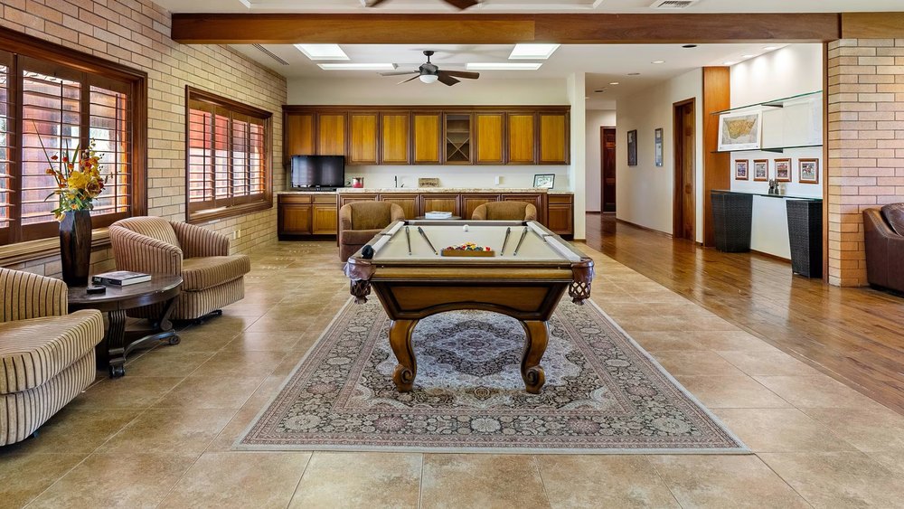 Spacious game room with brick accent walls, tiled flooring, a central billiards table on an ornate area rug, and built-in wood cabinetry in the background.