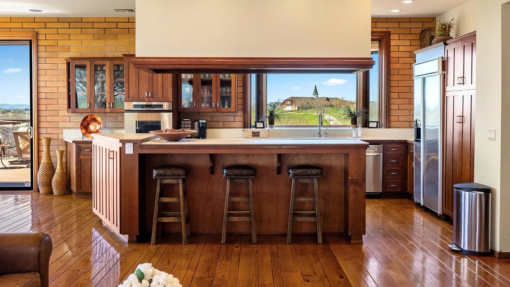 Spacious rustic kitchen featuring rich wood cabinetry, brick walls, a large center island with counter seating, and a large window framing scenic outdoor views.