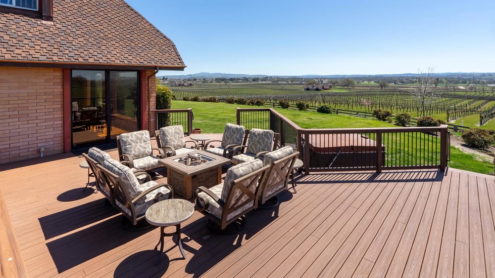 Outdoor wooden deck with plush cushioned armchairs centered around a square fire pit table, overlooking expansive vineyard fields stretching to distant hills.