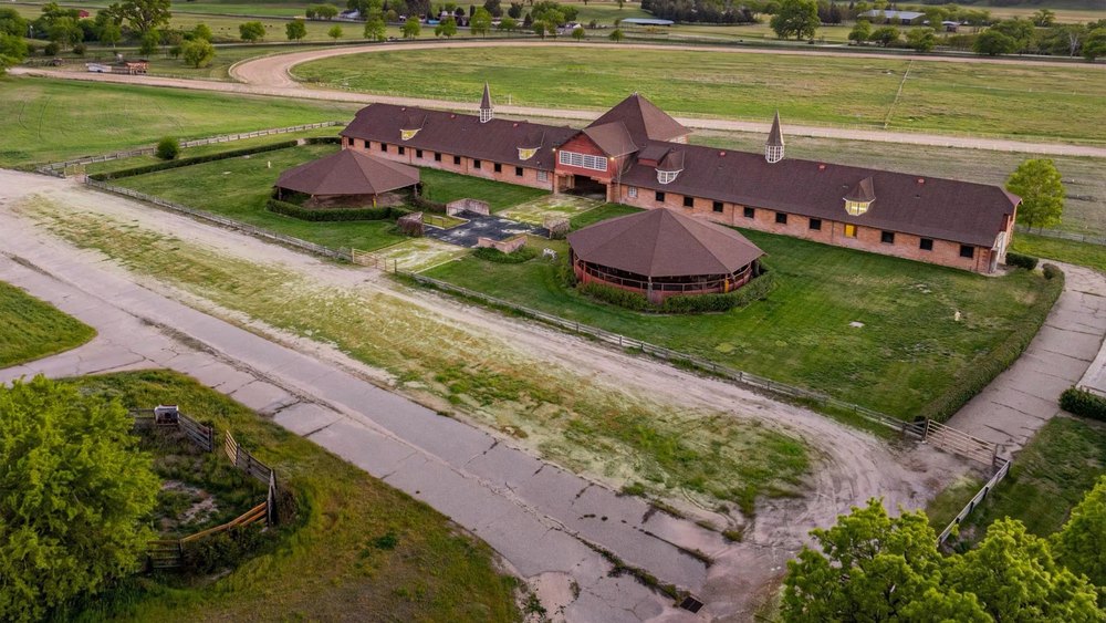 Aerial view of an equestrian facility featuring a long red-roofed barn structure, round covered arenas, and green pastures bordered by fencing.
