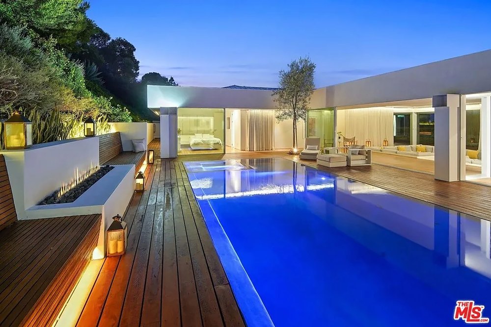 Modern backyard oasis at night with an illuminated pool, built-in fire pit, and wooden benches.